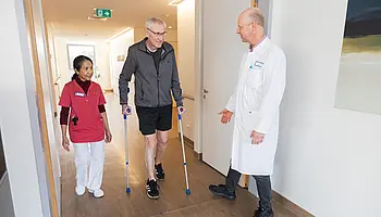 Ein älterer Mann geht mit zwei Unterarmgehstützen durch einen Klinikflur. Eine Pflegekraft begleitet ihn, ein Arzt steht ihm gegenüber und spricht mit ihm.