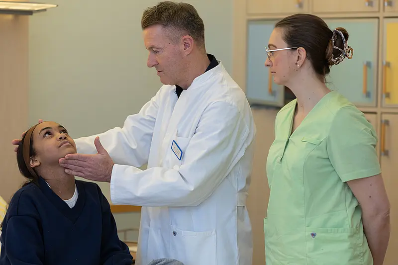 Dr. PD Daniel Windschall untersucht den Hals- und Kieferbereich eines sitzenden Kindes. Eine Pflegefachkraft steht daneben.