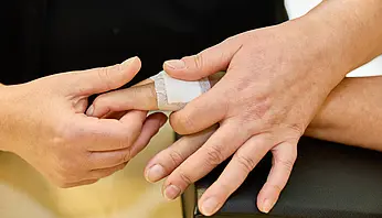 Nahaufnahme von Händen, während eine Ergotherapeutin einen Verband oder eine Schiene an einem Finger anlegt.