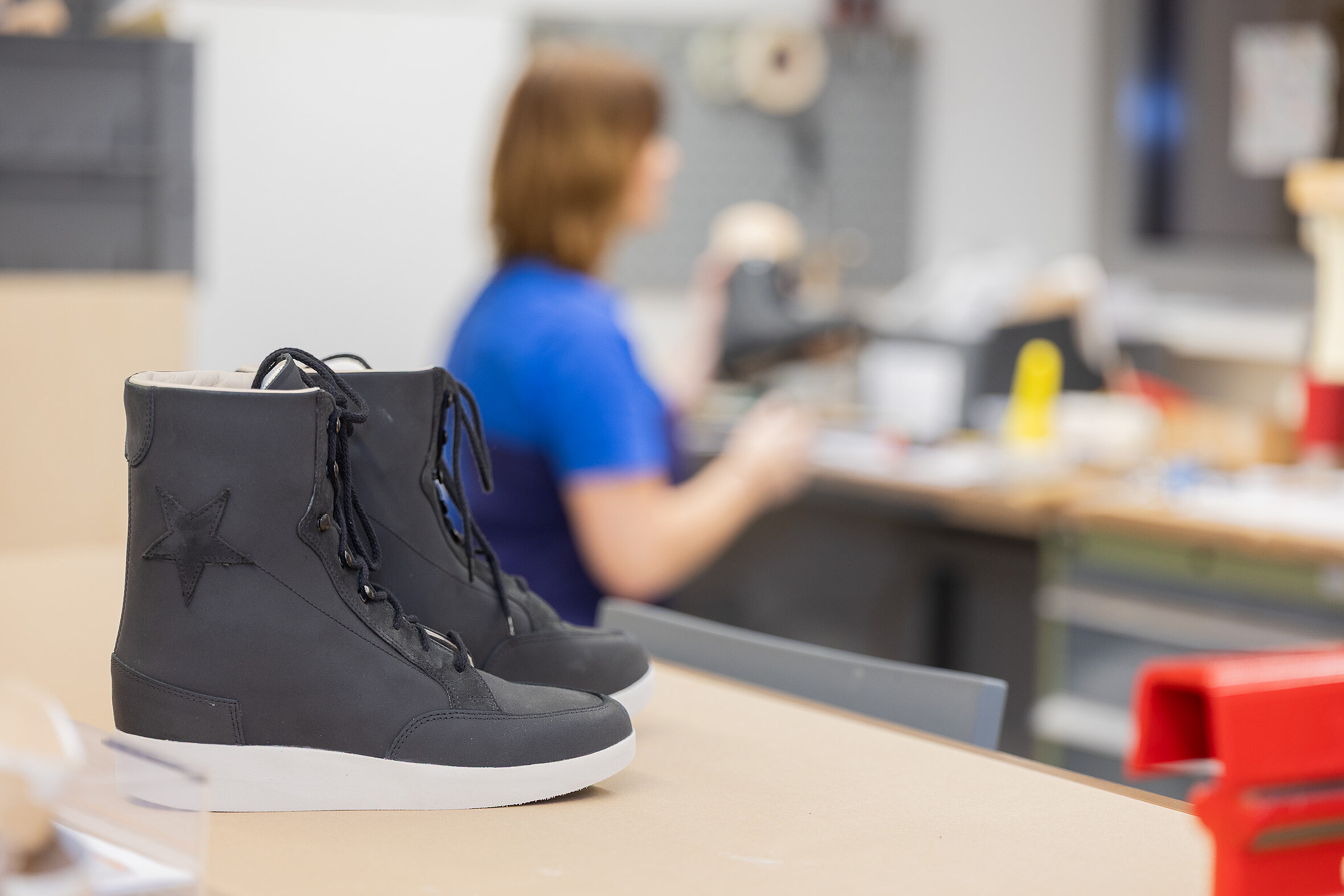 Ein schwarzer orthopädischer Schuh steht auf einer Werkbank, im Hintergrund ist unscharf eine Fachkraft bei der handwerklichen Arbeit zu sehen.