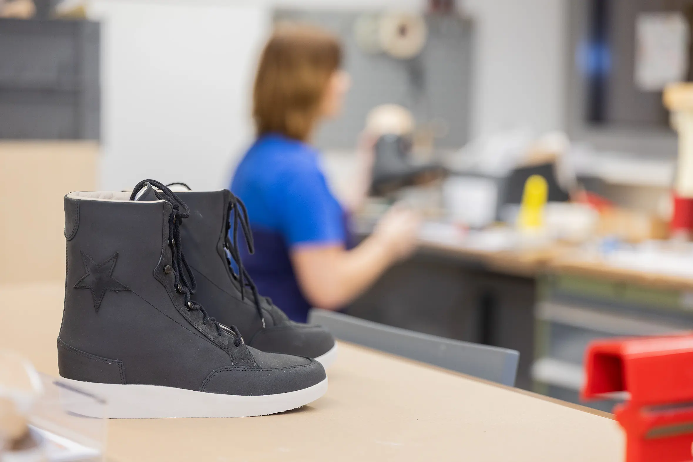 Ein schwarzer orthopädischer Schuh steht auf einer Werkbank, im Hintergrund ist unscharf eine Fachkraft bei der handwerklichen Arbeit zu sehen.