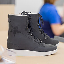 Ein schwarzer orthopädischer Schuh steht auf einer Werkbank, im Hintergrund ist unscharf eine Fachkraft bei der handwerklichen Arbeit zu sehen.