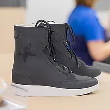 Ein schwarzer orthopädischer Schuh steht auf einer Werkbank, im Hintergrund ist unscharf eine Fachkraft bei der handwerklichen Arbeit zu sehen.