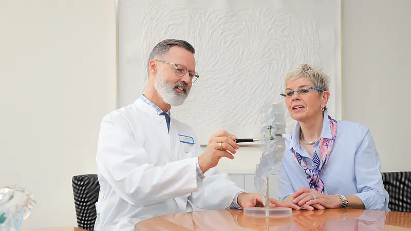 Chefarzt Dr. Brinkmann erklärt einer Patientin anhand eines transparenten Wirbelsäulenmodells den Aufbau der Wirbelsäule und das Vorgehen während der OP.