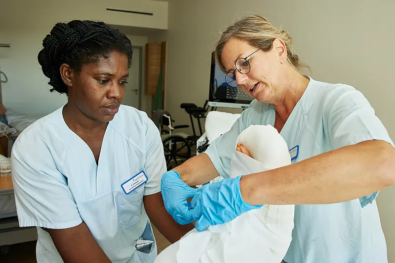 Zwei Pflegefachkräfte legen bei einer liegenden Patientin einen schützenden Verband an und stimmen die einzelnen Handgriffe miteinander ab.
