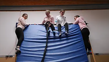 Jugendliche sitzen und bewegen sich gemeinsam auf einer großen schrägen Weichbodenmatte.