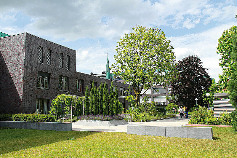 Grünanlage mit Wegen, Bäumen und modernen Gebäuden im Hintergrund; mehrere Personen gehen spazieren.