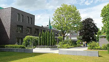 Grünanlage mit Wegen, Bäumen und modernen Gebäuden im Hintergrund; mehrere Personen gehen spazieren.