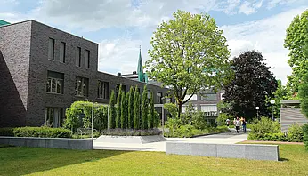 Grünanlage mit Wegen, Bäumen und modernen Gebäuden im Hintergrund; mehrere Personen gehen spazieren.