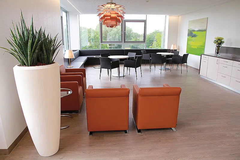 Runder Tisch mit Stühlen vor einem großformatigen Wandbild in Grüntönen, Tageslicht durch Fenster.