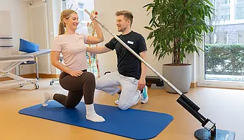 Eine Patientin führt auf einer Matte eine Übung im Ausfallschritt mit einer Trainingsstange aus, während ein Physiotherapeut sie anleitet und korrigiert.