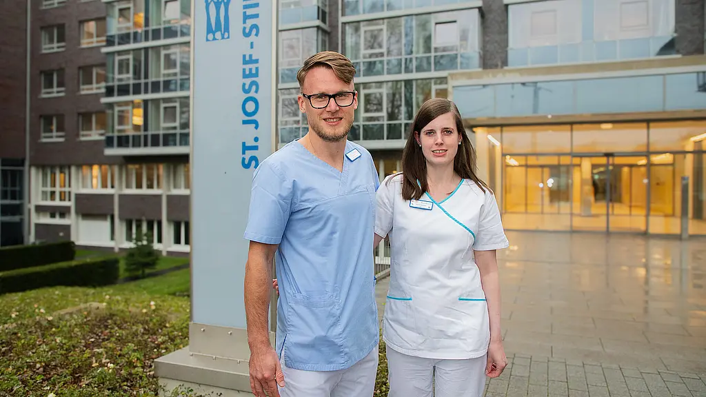 Ein Mann und eine Frau stehen vor einem warm beleuchteten Krankenhauseingang, hinter ihnen ein Krankenhausschild.