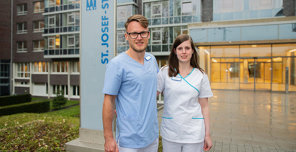 Ein Mann und eine Frau stehen vor einem warm beleuchteten Krankenhauseingang, hinter ihnen ein Krankenhausschild.