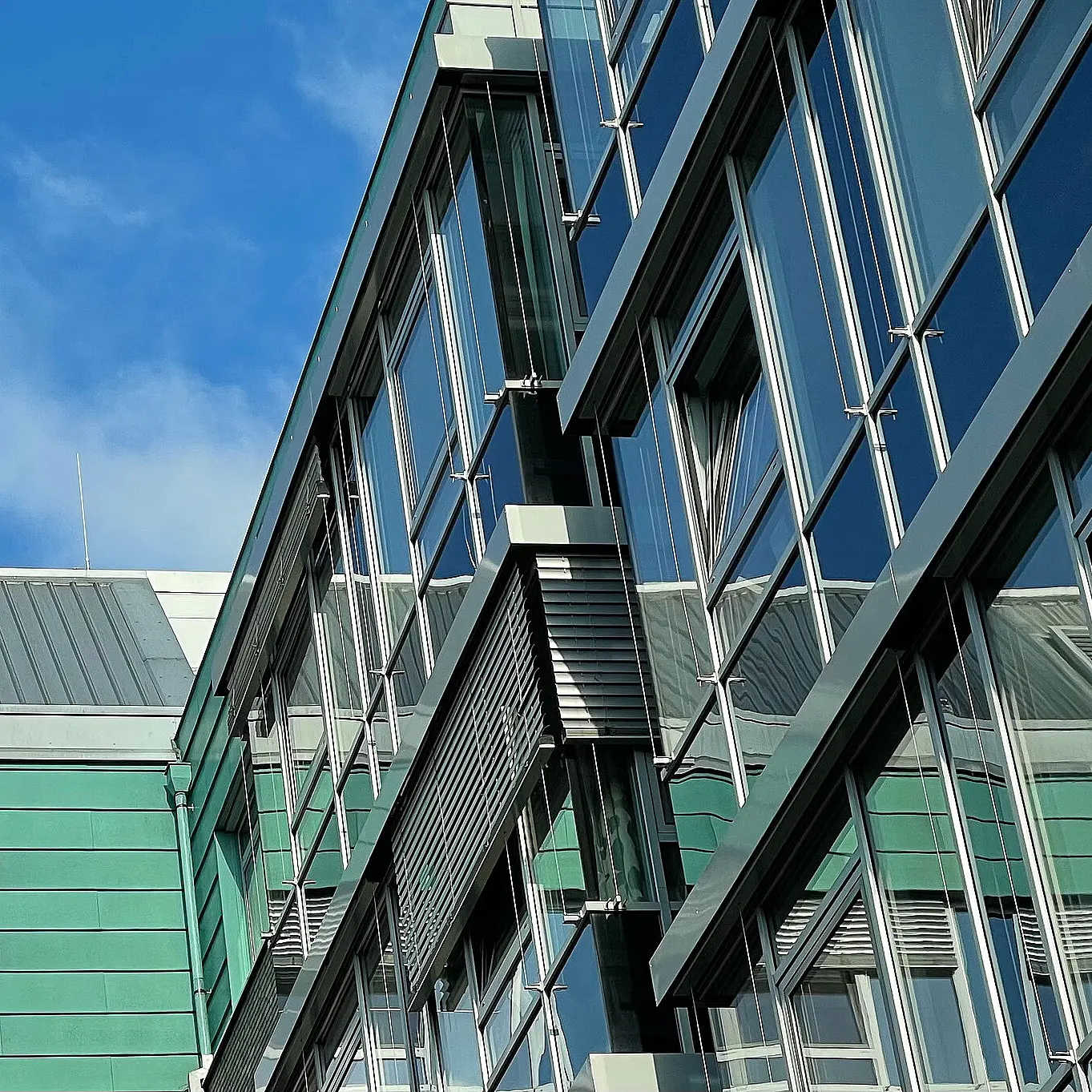 Schräg aufgenommene Ansicht einer modernen Glas- und Metallfassade eines Gebäudes des St. Josef-Stift Sendenhorst vor blauem Himmel.