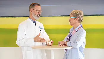 Chefarzt Dr. Brinkmann und Patientin stehen an einem Tisch und betrachten gemeinsam ein kleines Wirbelsäulenmodell.