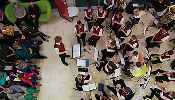 Blasorchester mit verschiedenen Instrumenten spielt in einem Innenraum; die Musikerinnen und Musiker sitzen im Halbkreis, Publikum ist anwesend.