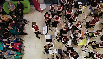 Blasorchester mit verschiedenen Instrumenten spielt in einem Innenraum; die Musikerinnen und Musiker sitzen im Halbkreis, Publikum ist anwesend.