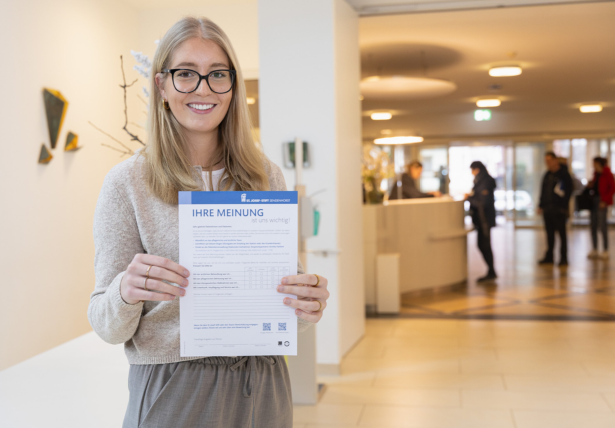 Mitarbeiterin im Eingangsbereich präsentiert einen Feedbackbogen zur Zufriedenheit mit Behandlung, Pflege, Therapie und Service.