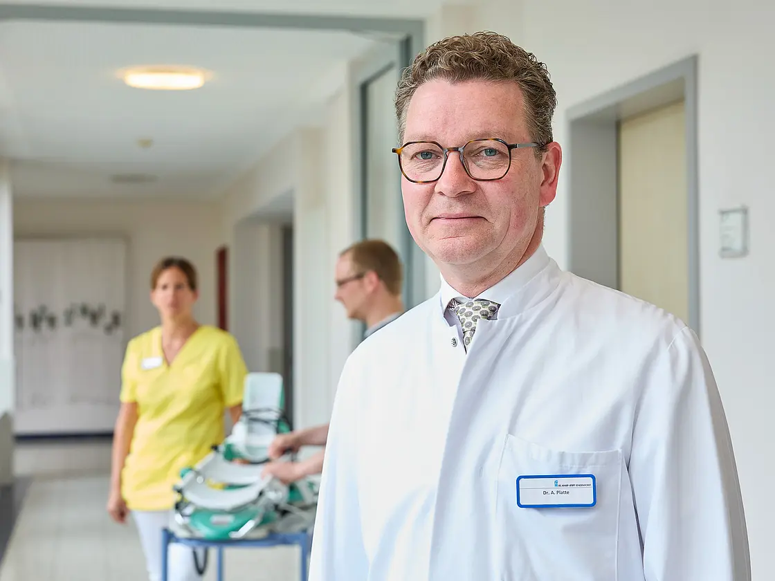 Ein Arzt im weißen Kittel steht im Flur einer Klinik; im Hintergrund bewegt sich Pflegepersonal mit einem Gerätewagen.
