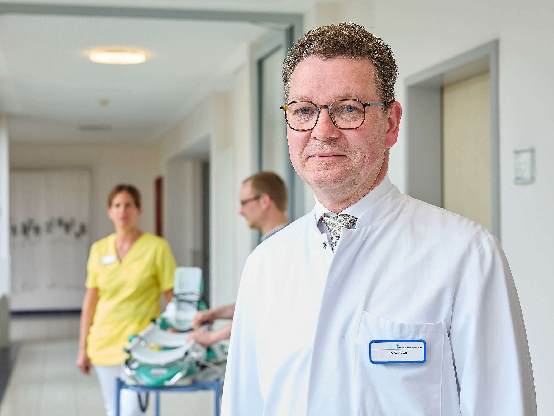 Ein Arzt im weißen Kittel steht im Flur einer Klinik; im Hintergrund bewegt sich Pflegepersonal mit einem Gerätewagen.