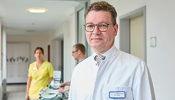 Ein Arzt im weißen Kittel steht im Flur einer Klinik; im Hintergrund bewegt sich Pflegepersonal mit einem Gerätewagen.