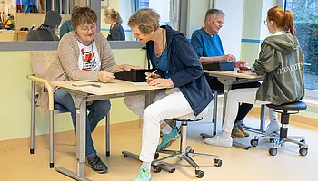 Therapieraum mit zwei Arbeitsplätzen im Vordergrund und einem weiteren Raum im Hintergrund: Eine Therapeutin begleitet einen erwachsene Patientin bei einer feinmotorischen Übung, während im selben Raum ein weiterer Erwachsener therapeutisch behandelt wird.