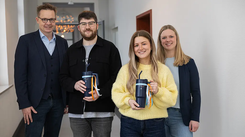 Vier Menschen stehen in einem Flur nebeneinander und lächeln, die beiden Auszubildenden in der Mitte halten jeweils einen blauen Trinkbecher in der Hand.