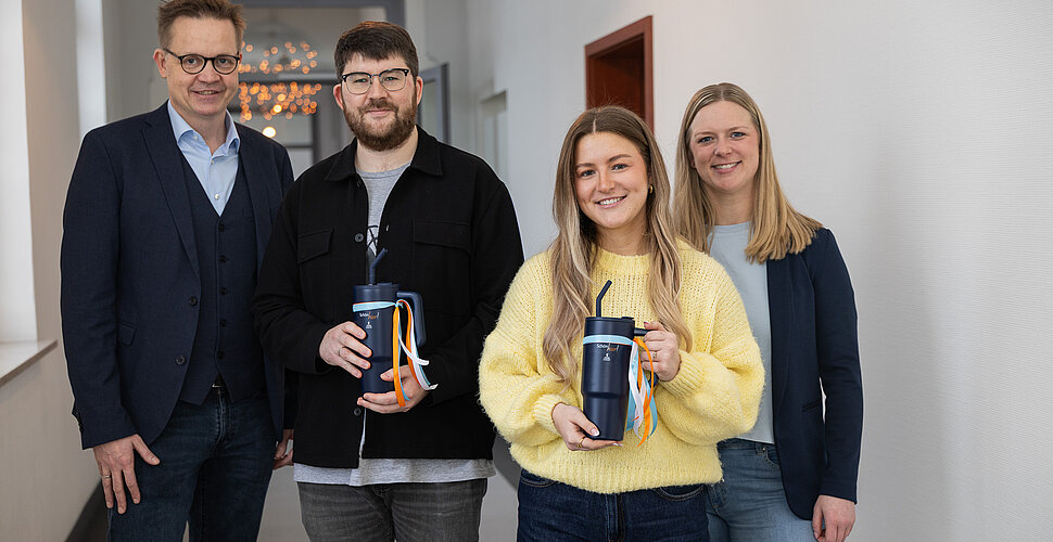 „Schön hier!“ – das gilt auch nach der Ausbildung: Thomas Lasthaus und Jule Neumann gratulieren Vorsitzender der Geschäftsführung Dr. Ansgar Klemann (l.) und Ausbildungskoordinatorin Laura Niesmann (r.) zum Ausbildungsabschluss. Vier Menschen stehen in einem Flur nebeneinander und lächeln, die beiden Auszubildenden in der Mitte halten jeweils einen blauen Trinkbecher in der Hand.