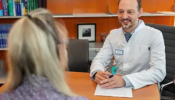 PD Dr. med. Peter Korsten sitzt an einem Schreibtisch und spricht mit einer Patientin, im Hintergrund stehen Akten und Bücherregale.