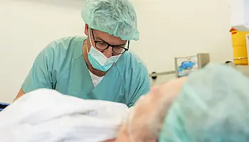 Anästhesist in OP-Kleidung überwacht einen liegenden Patienten und kontrolliert Zugänge und Schläuche am OP-Tisch.