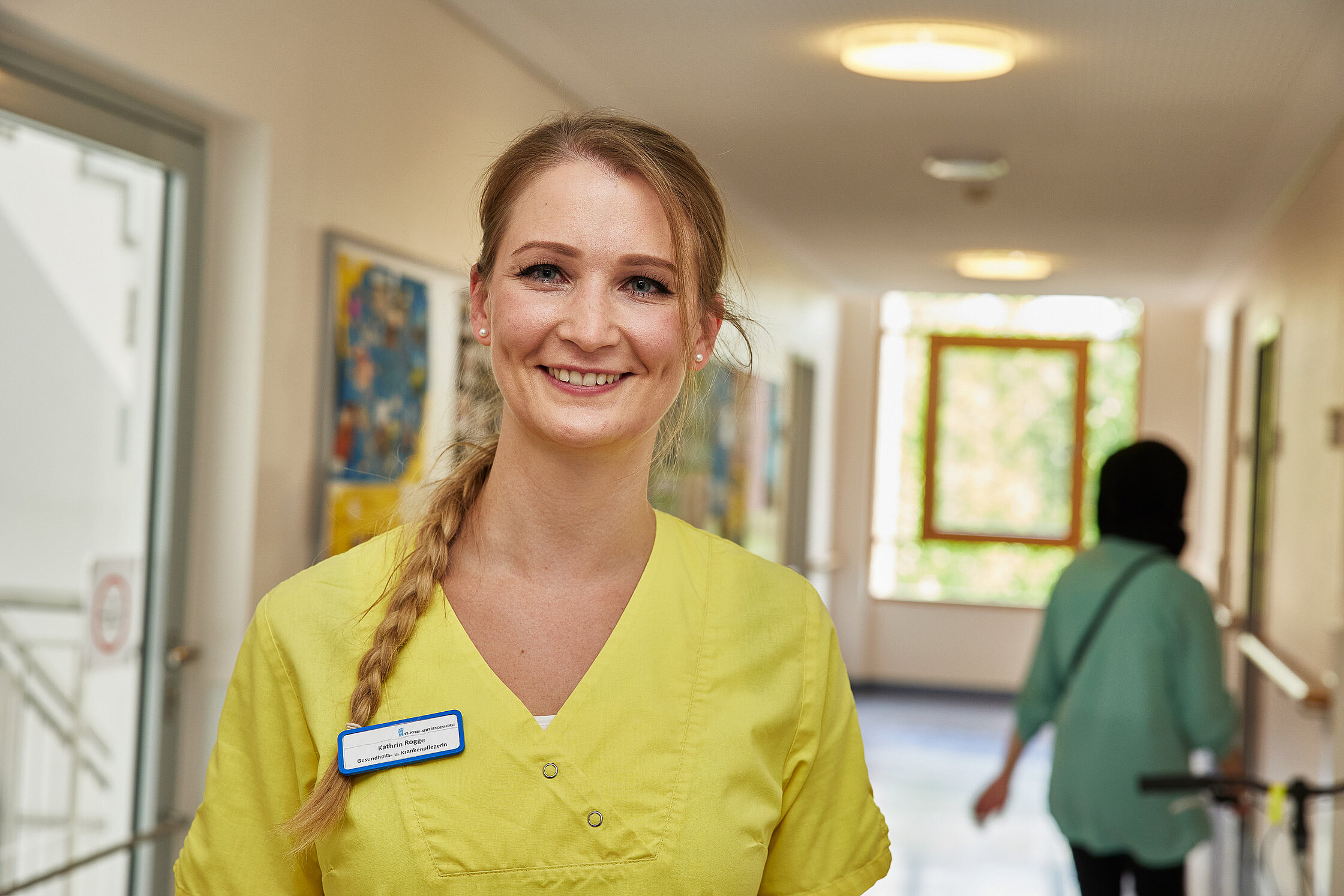 Eine Pflegefachkraft steht im Flur der Station und blickt freundlich in die Kamera, im Hintergrund bewegt sich eine weitere Person.
