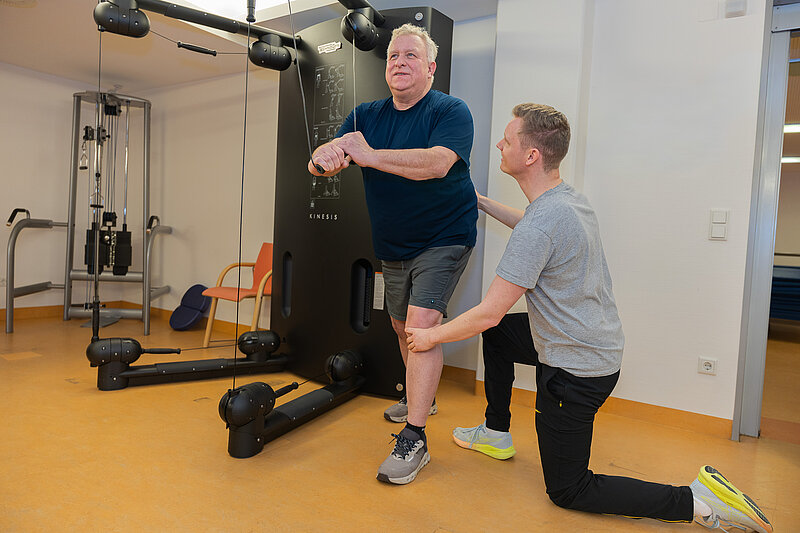 Ein Patient trainiert an einem Seilzuggerät, während ein Therapeut kniend das Knie stabilisiert.