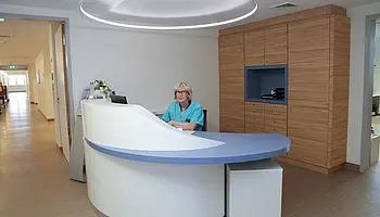 Heller Stationsflur mit geschwungenem Empfangstresen; eine Pflegekraft sitzt am Arbeitsplatz, im Hintergrund angrenzende Räume.