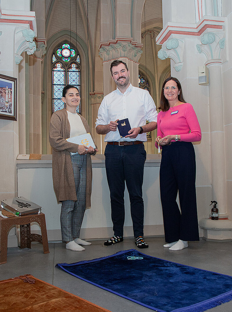 Drei Personen stehen barfuß in einem hohen Kapellenraum mit Säulen und Buntglasfenstern; vor ihnen liegen unterschiedlich farbige Gebetsteppiche auf dem Boden, eine Person hält ein Buch in den Händen.