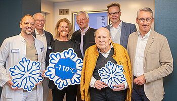 Mehrere Erwachsene stehen in einem Eingangsbereich zusammen und halten Schneeflocken-Symbole mit der Aufschrift „−110 °C“ in den Händen.