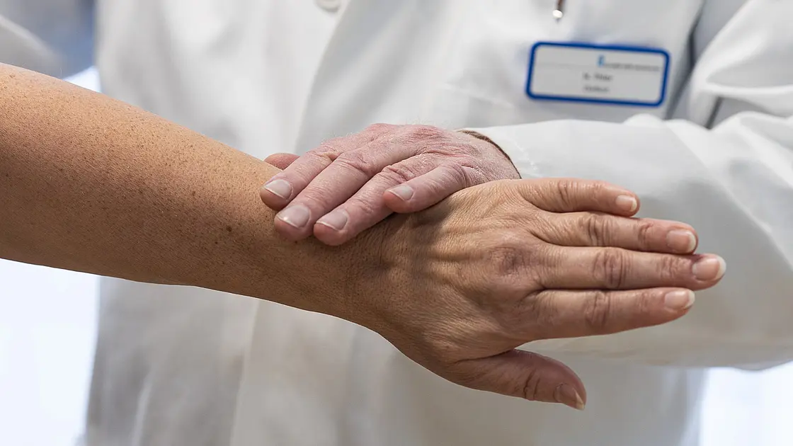 Nahaufnahme von zwei Händen: Eine ärztliche Hand liegt auf dem Unterarm einer Patientin während einer Untersuchung.