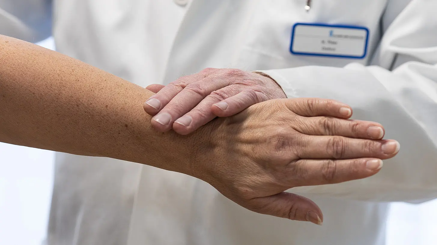 Nahaufnahme von zwei Händen: Eine ärztliche Hand liegt auf dem Unterarm einer Patientin während einer Untersuchung.