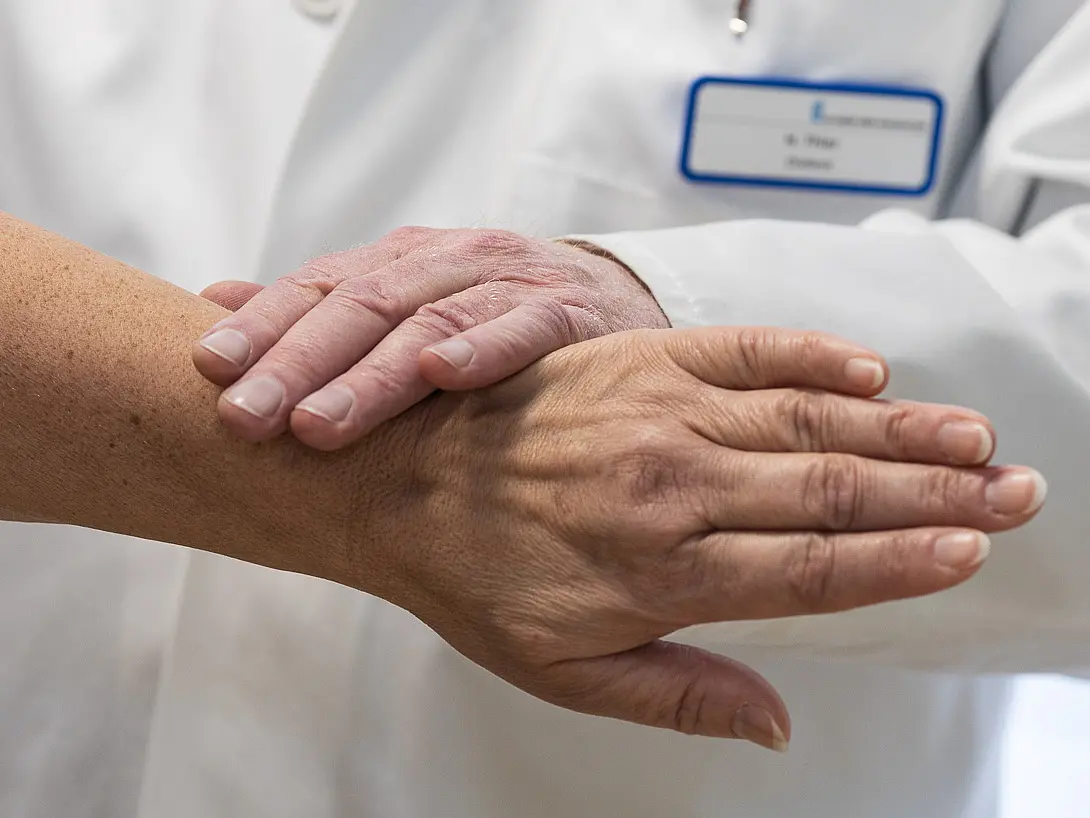 Nahaufnahme von zwei Händen: Eine ärztliche Hand liegt auf dem Unterarm einer Patientin während einer Untersuchung.