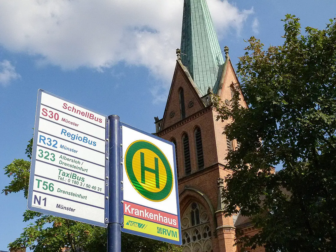 Haltestellenschild mit Linienangaben für Schnellbus, RegioBus und TaxiBus, im Hintergrund ein Kirchengebäude auf dem Gelände des St. Josef-Stift Sendenhorst.