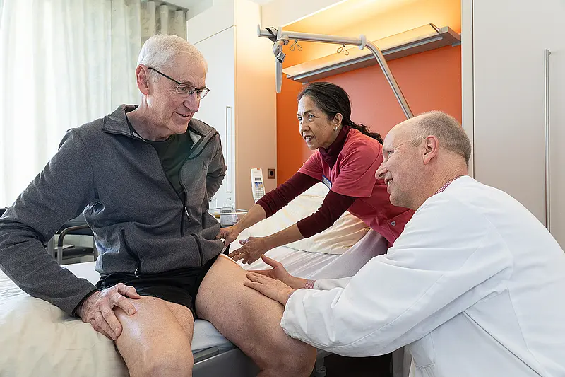 Ein Patient sitzt auf dem Bett, während eine Pflegekraft und ein Arzt neben ihm stehen und sein Bein untersuchen. Alle drei sind im Gespräch.