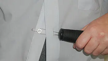 Nahaufnahme einer Hand, die mithilfe einer Knöpfhilfe einen Hemdknopf durch das Knopfloch zieht; das Hilfsmittel unterstützt Menschen mit eingeschränkter Handfunktion beim selbstständigen An- und Auskleiden und schont die Gelenke.