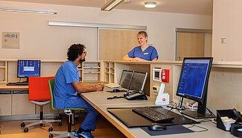Zwei Mitarbeitende in Dienstkleidung stehen und sitzen an einem zentralen Tresen mit Monitoren und Telefonen.