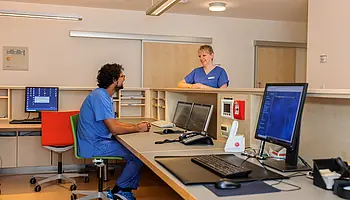Zwei Mitarbeitende in Dienstkleidung stehen und sitzen an einem zentralen Tresen mit Monitoren und Telefonen.