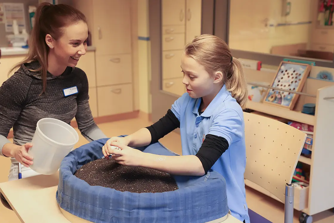 Ergotherapeutische Fachkraft begleitet ein Mädchen mit Kinderrheuma bei einer beidhändigen Übung in einer mit Material gefüllten Schale an einem Tisch.