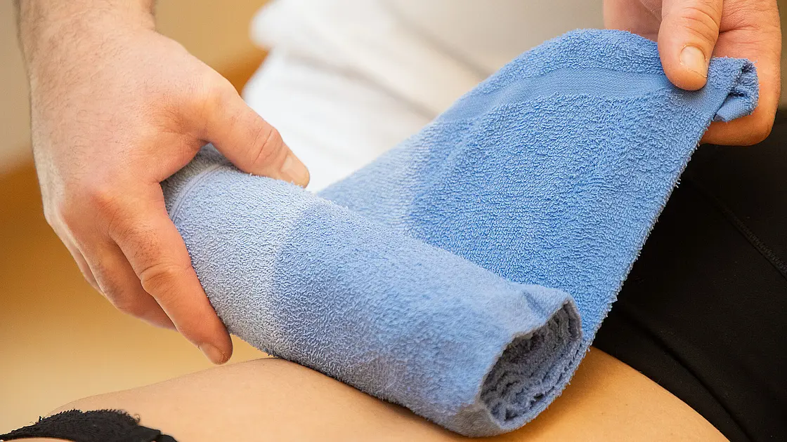 Eine Fachkraft rollt ein blaues, feucht-warmes Handtuch auf dem Oberschenkel einer Patientin oder eines Patienten ab. Die Behandlung findet auf einer Therapieliege statt.