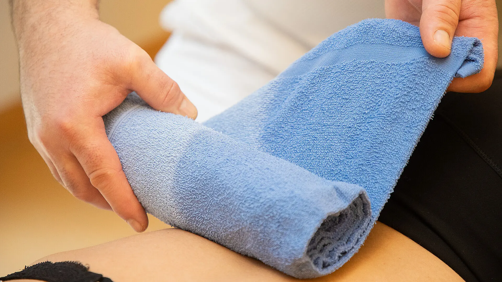 Eine Fachkraft rollt ein blaues, feucht-warmes Handtuch auf dem Oberschenkel einer Patientin oder eines Patienten ab. Die Behandlung findet auf einer Therapieliege statt.