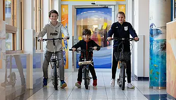 Drei Kinder fahren mit Laufrädern durch einen hellen Klinikflur mit farbigen Wandmotiven.
