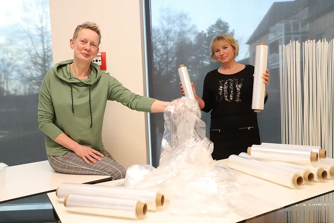 Zwei Mitarbeitende präsentieren Folienrollen zum Verpacken von Speisen und Speisentransportwagen.