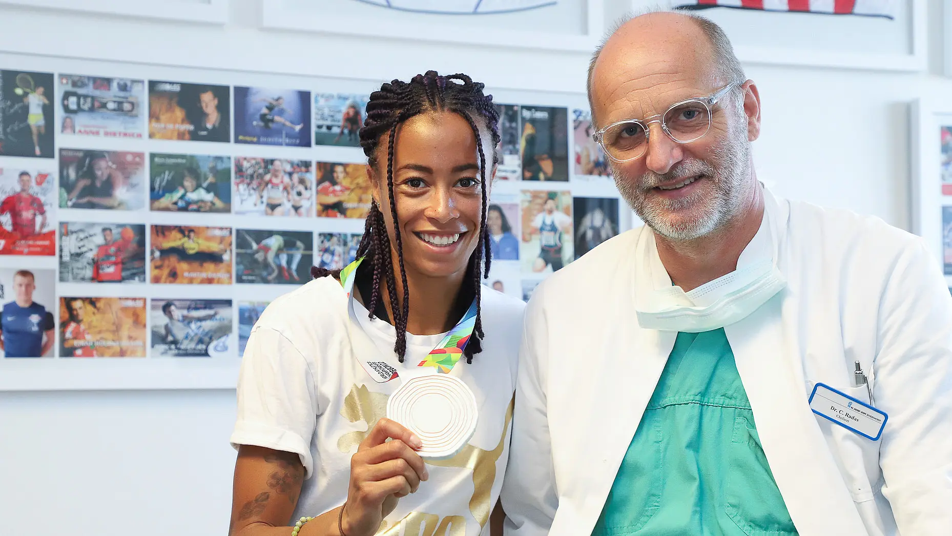 Die Leichtathletin Tatjana Pinto steht neben Chefarzt Dr. Carsten Radas und hält ihre Bronzemedaille der Weltmeisterschaft in die Kamera. Beide lächeln.