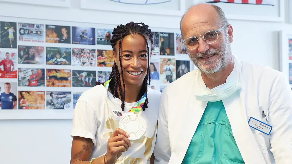 Die Leichtathletin Tatjana Pinto steht neben Chefarzt Dr. Carsten Radas und hält ihre Bronzemedaille der Weltmeisterschaft in die Kamera. Beide lächeln.